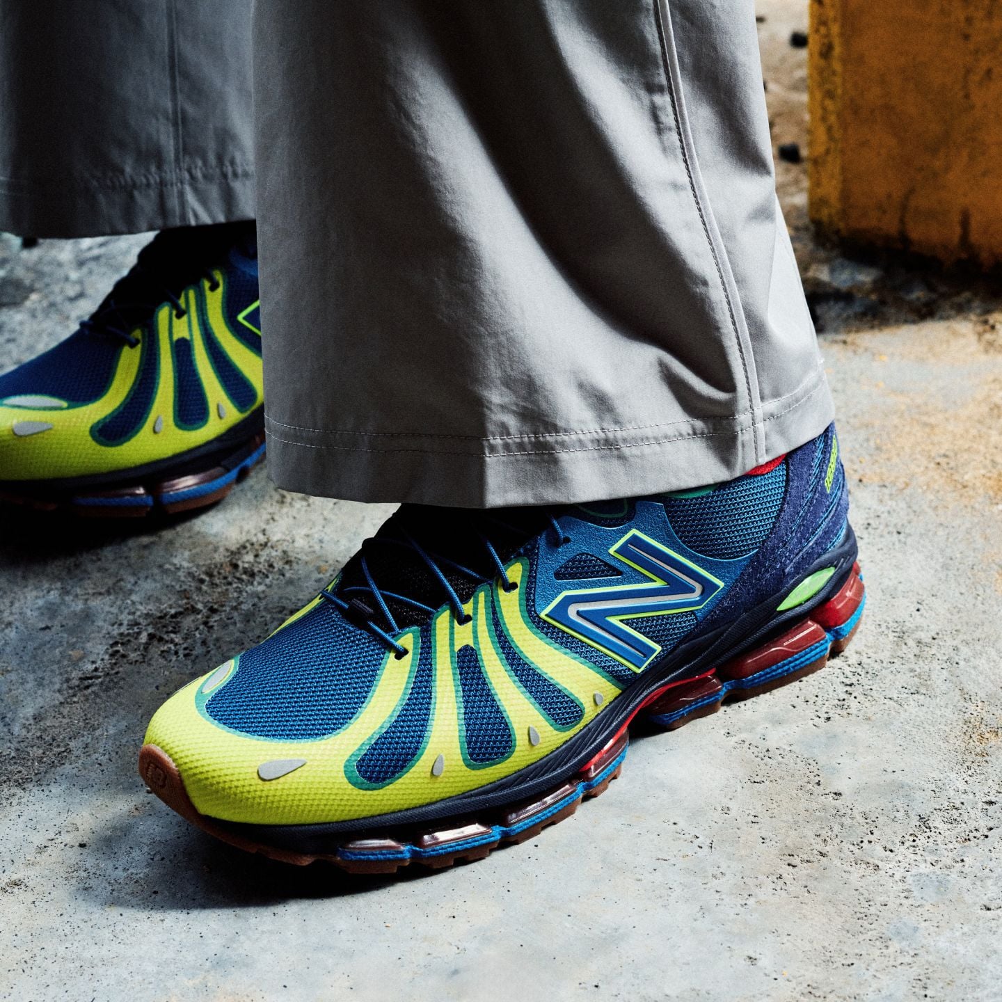 Close-up of someone wearing blue and green New Balance sneakers on a concrete background. 