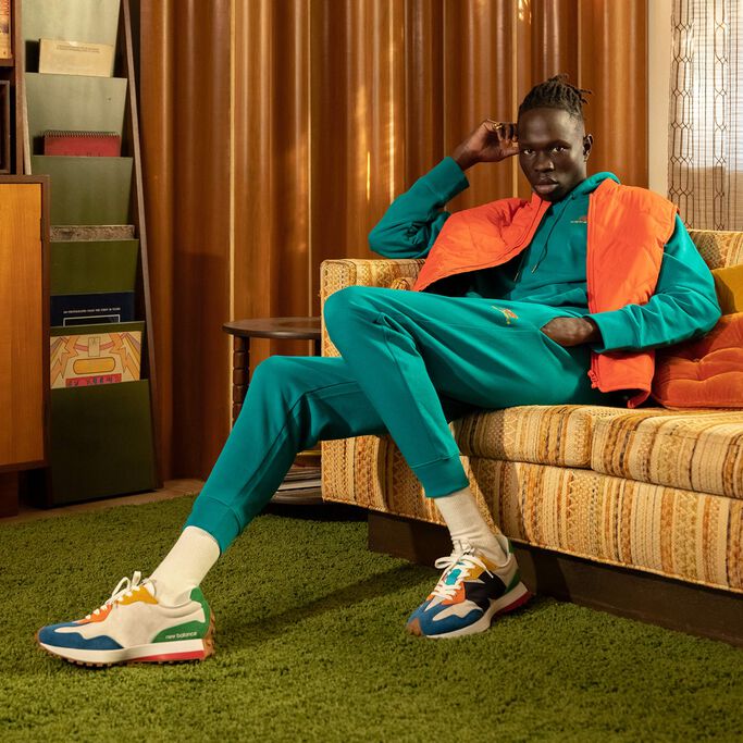A man wearing teal sweatpants and multicolor NB 327 sneakers sitting on a couch in a 1970s décor room