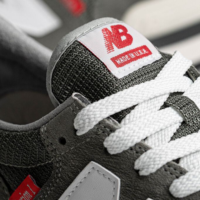 Detail crop of the tongue and upper of a grey NB 990 Version 1 sneaker accented with red labels