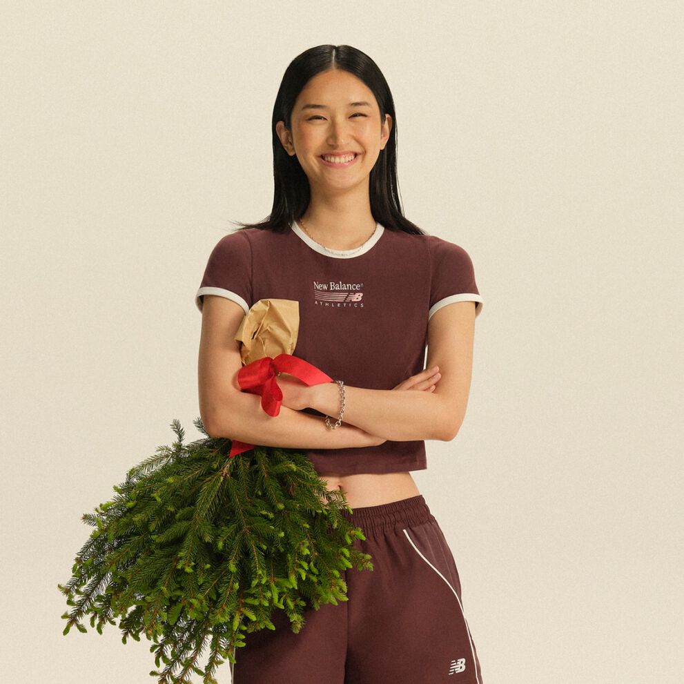 Person wearing a burgundy New Balance athletic set holding a wrapped gift and evergreen branches against a neutral studio background. 