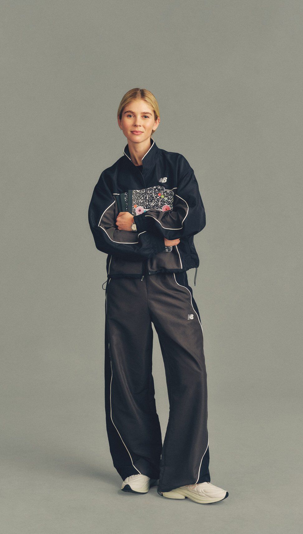 Full-body studio image of a person in a black New Balance tracksuit holding a patterned notebook against a neutral gray background.