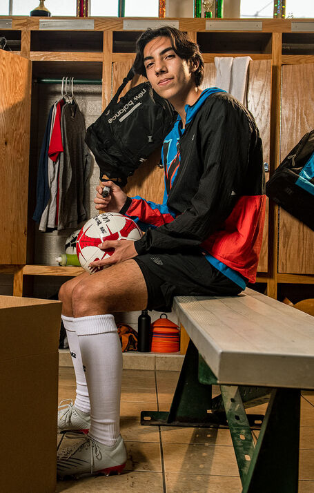 A teenage boy sitting in a football locker room wearing an NB Impact Run Light Pack Jacket as he writes on a football to send to professional footballer and NB athlete, Sadio Mané