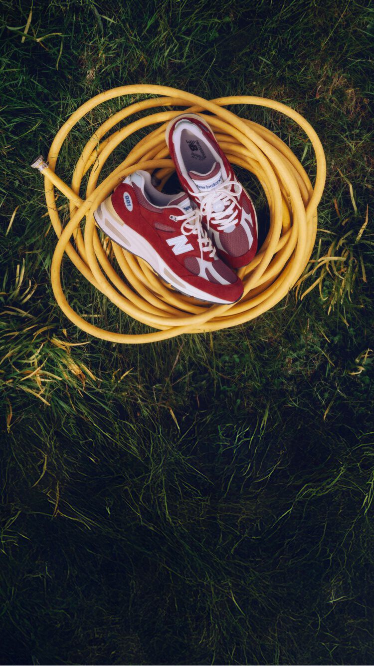 Une paire d'espadrilles New Balance Made in UK pos&eacute;es sur un tuyau jaune enroul&eacute;, au milieu d'une pelouse verdoyante.&nbsp;