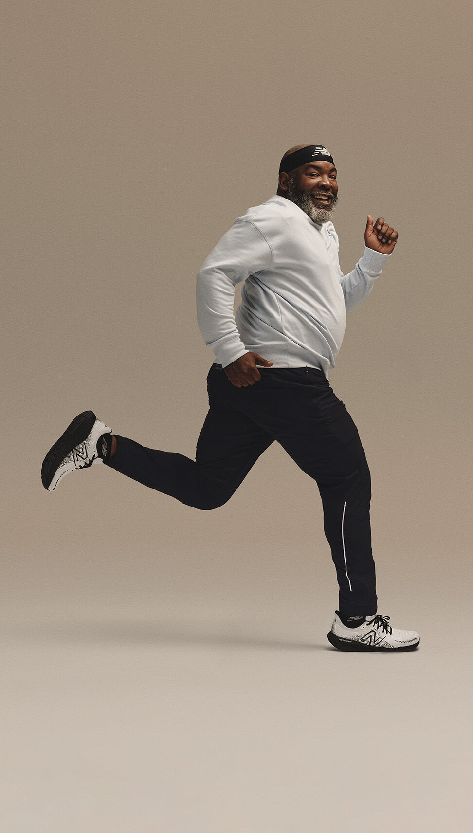 Homme d'âge moyen dans une pose de course devant un fond de studio beige portant de la tête aux pieds un équipement de course NB 