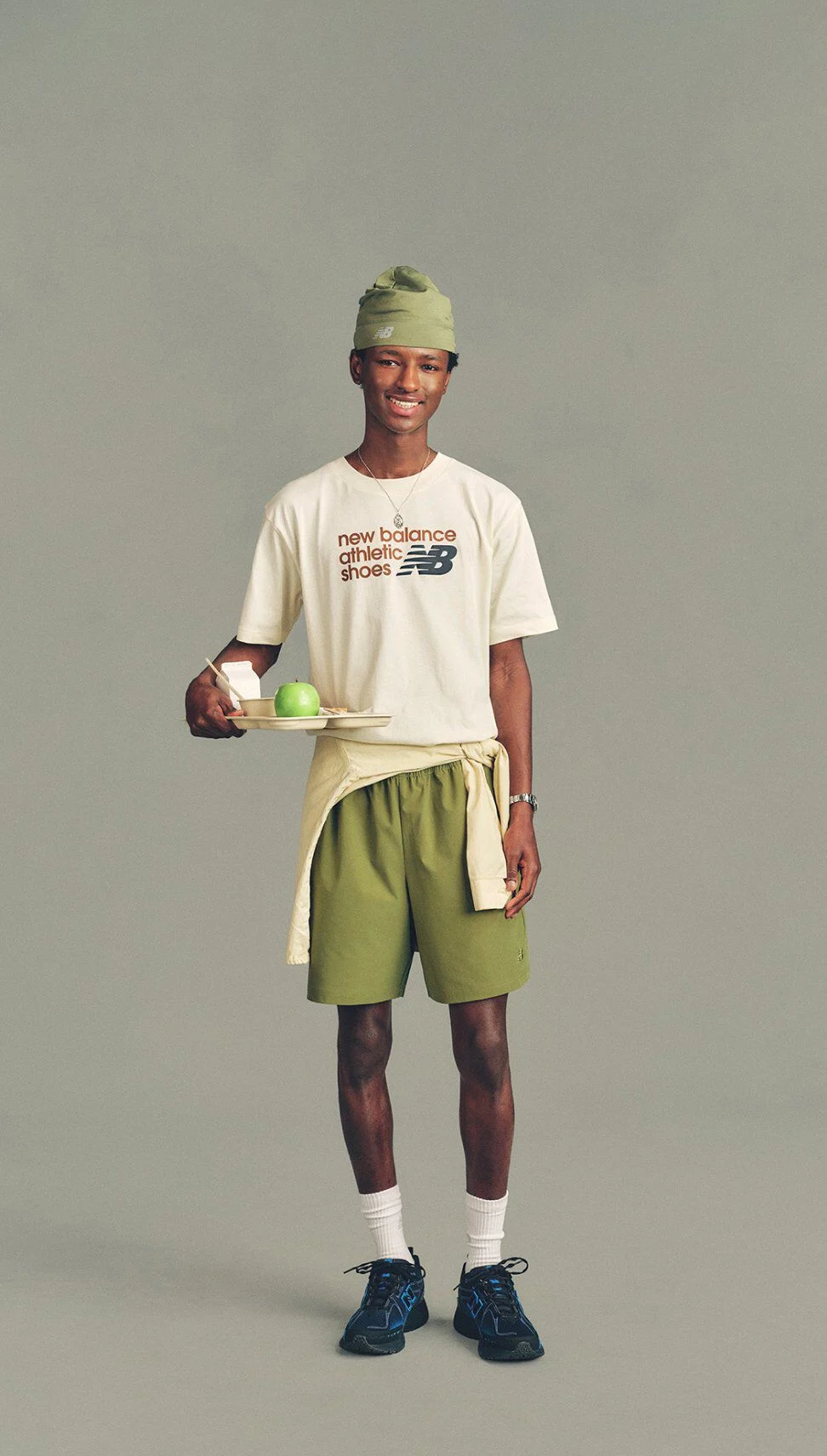 The person is wearing a white T-shirt with printed text reading &ldquo;new balance athletic shoes&rdquo; alongside the New Balance logo, paired with olive-green athletic shorts.