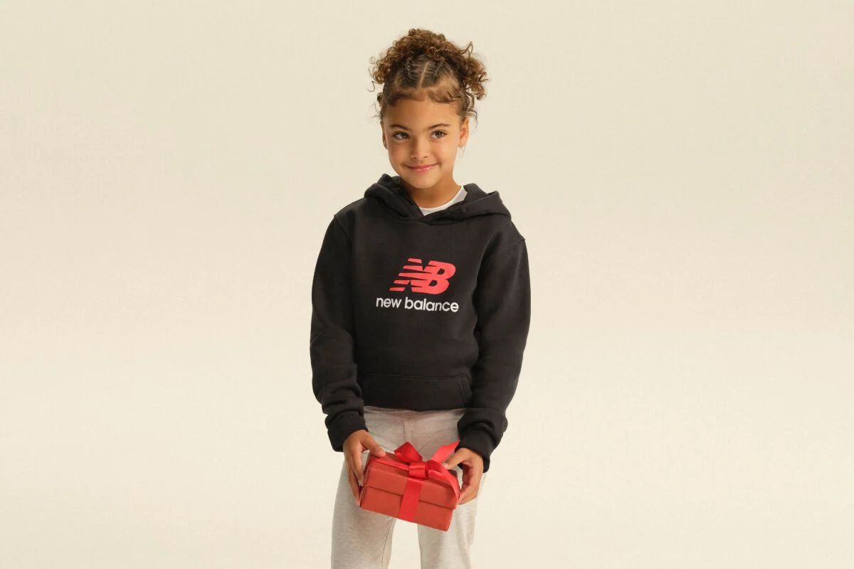 Child wearing a black New Balance hoodie and gray pants holding a red gift box against a light studio backdrop. 
