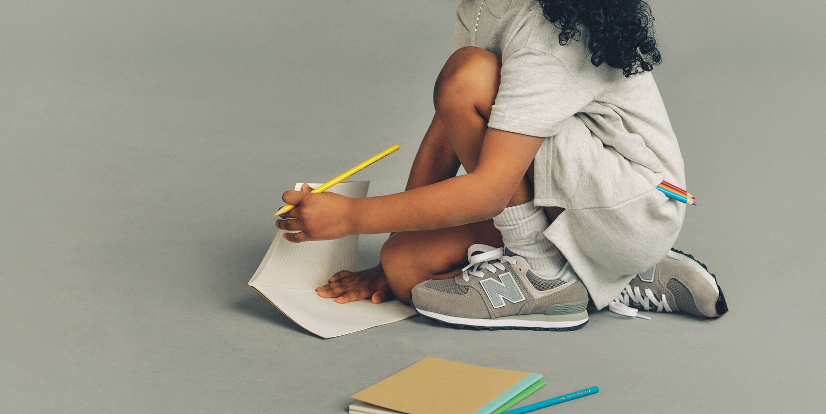 Gros plan pris en studio d'un enfant accroupi sur le sol, devant un fond uni gris clair, concentr&eacute; sur un dessin qu'il r&eacute;alise sur une feuille de papier lign&eacute;. 