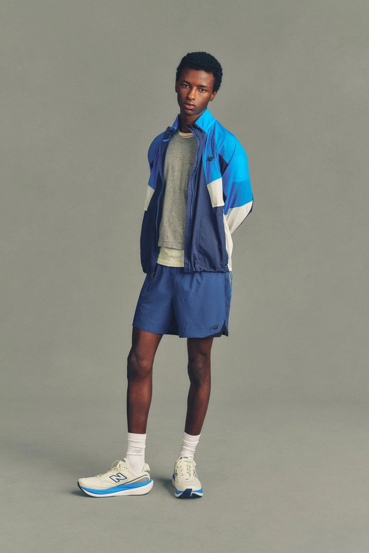 A man wearing a New Balance jacket, shirt and shorts standing in front of a grey studio background.