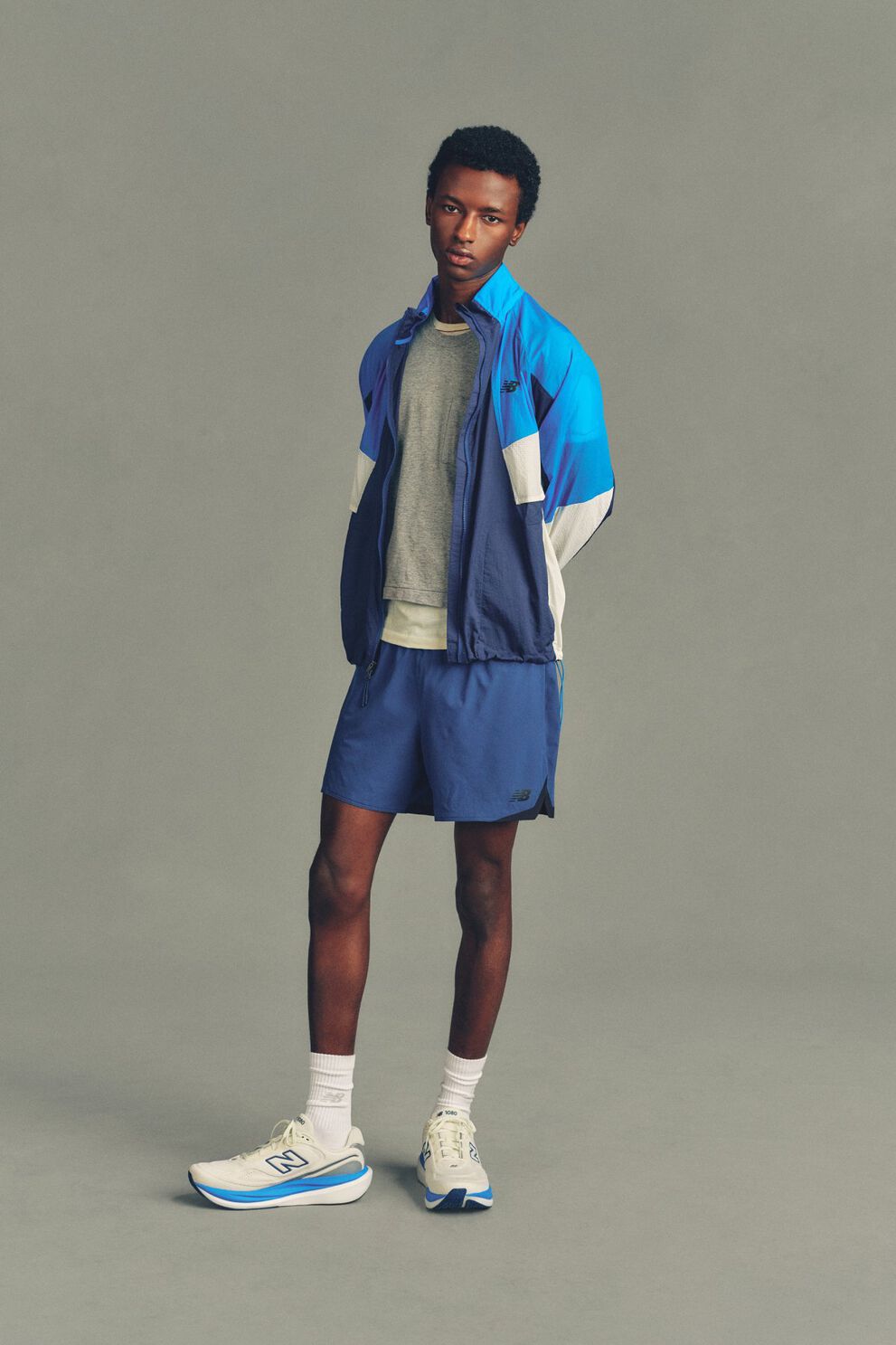 A man wearing a New Balance jacket, shirt and shorts standing in front of a grey studio background.