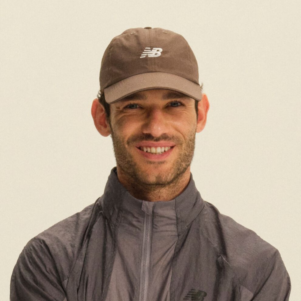 Person wearing a brown New Balance cap and a gray color-block performance jacket in a clean studio setting.