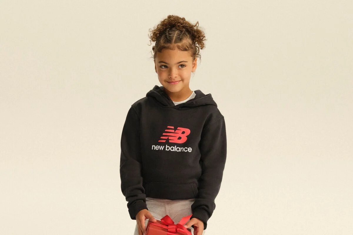 Enfant portant un chandail &agrave; capuchon New Balance noir et un pantalon d&rsquo;entra&icirc;nement gris, et tenant une bo&icirc;te-cadeau rouge dans un d&eacute;cor de studio &eacute;pur&eacute;.