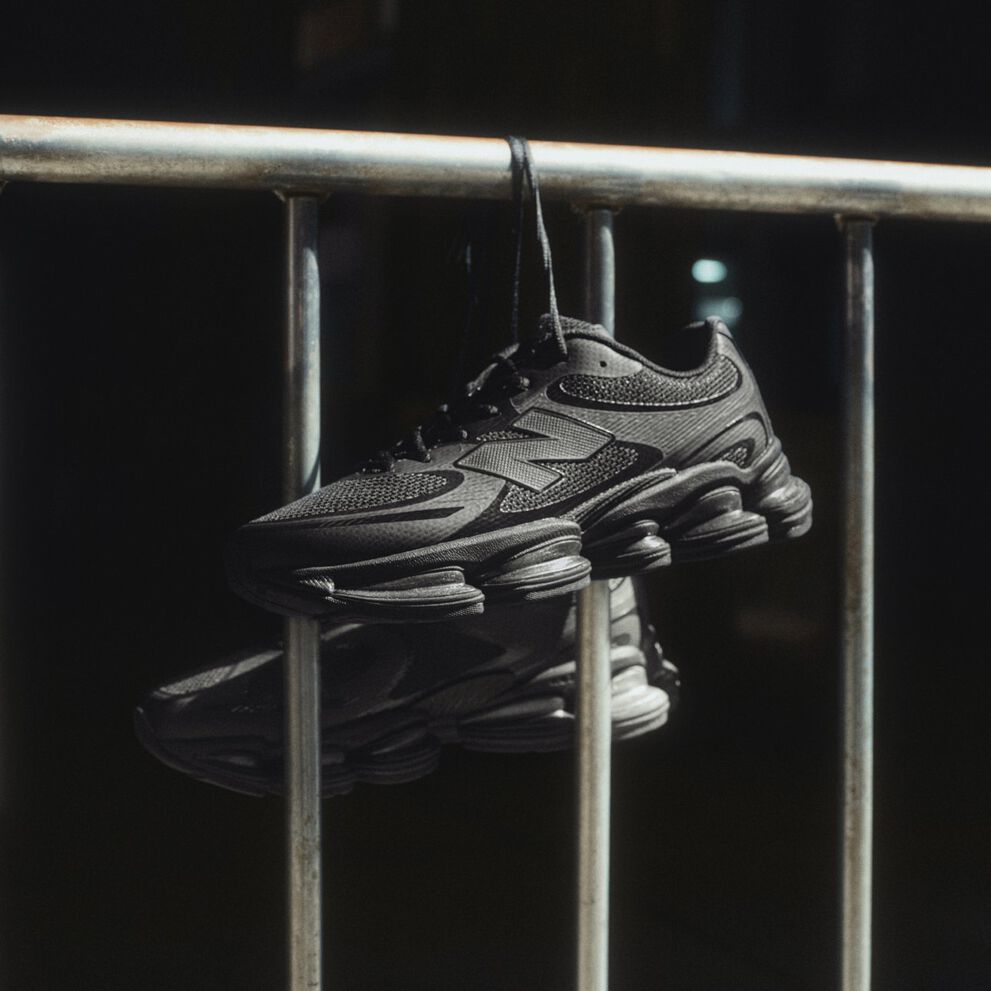 Baskets New Balance noires suspendues &agrave; des lacets sur une rambarde m&eacute;tallique sous un &eacute;clairage dramatique.