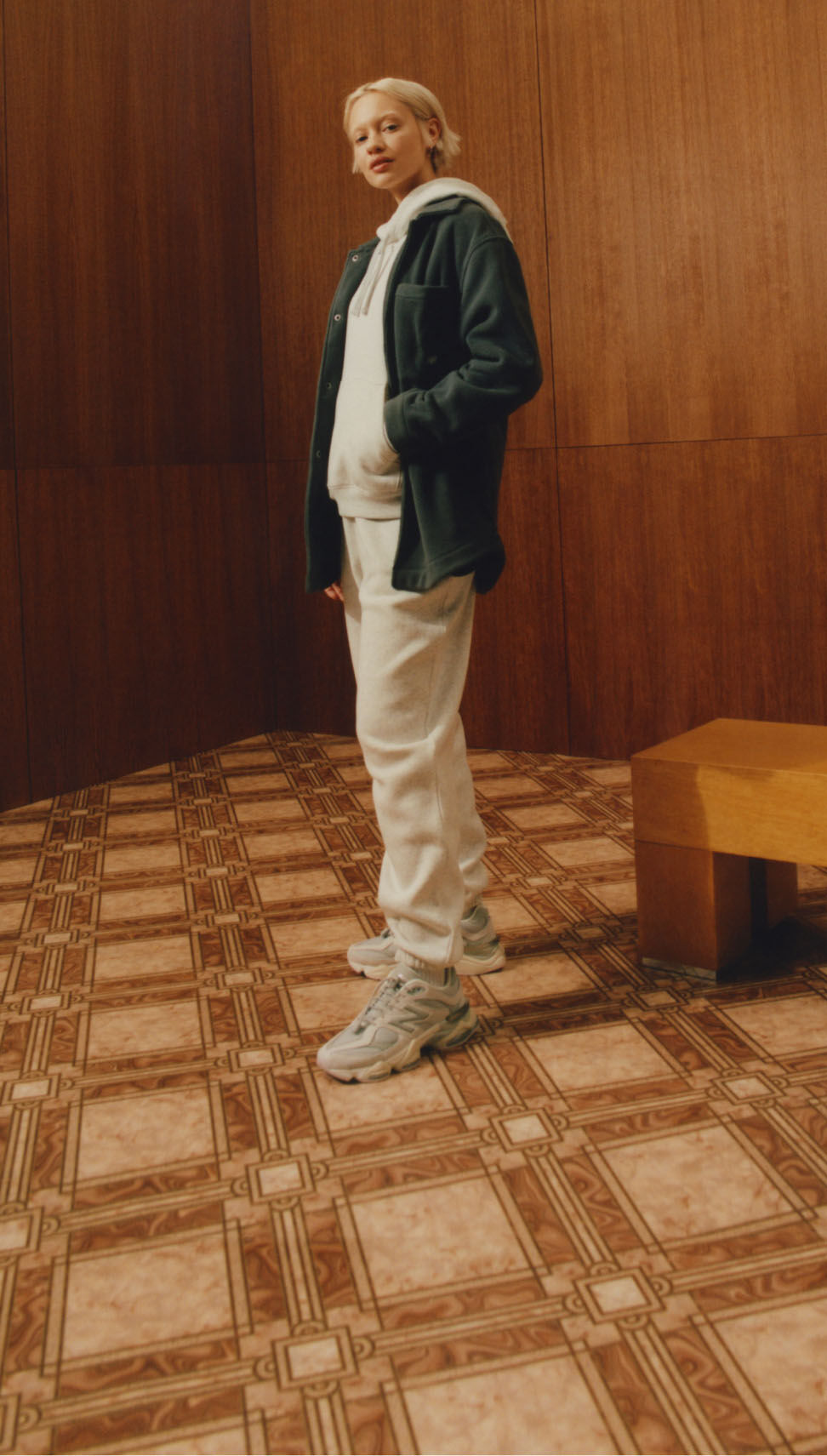 Une personne debout dans un environnement vintage en bois et tuiles, portant des vêtements et des chaussures New Balance. 