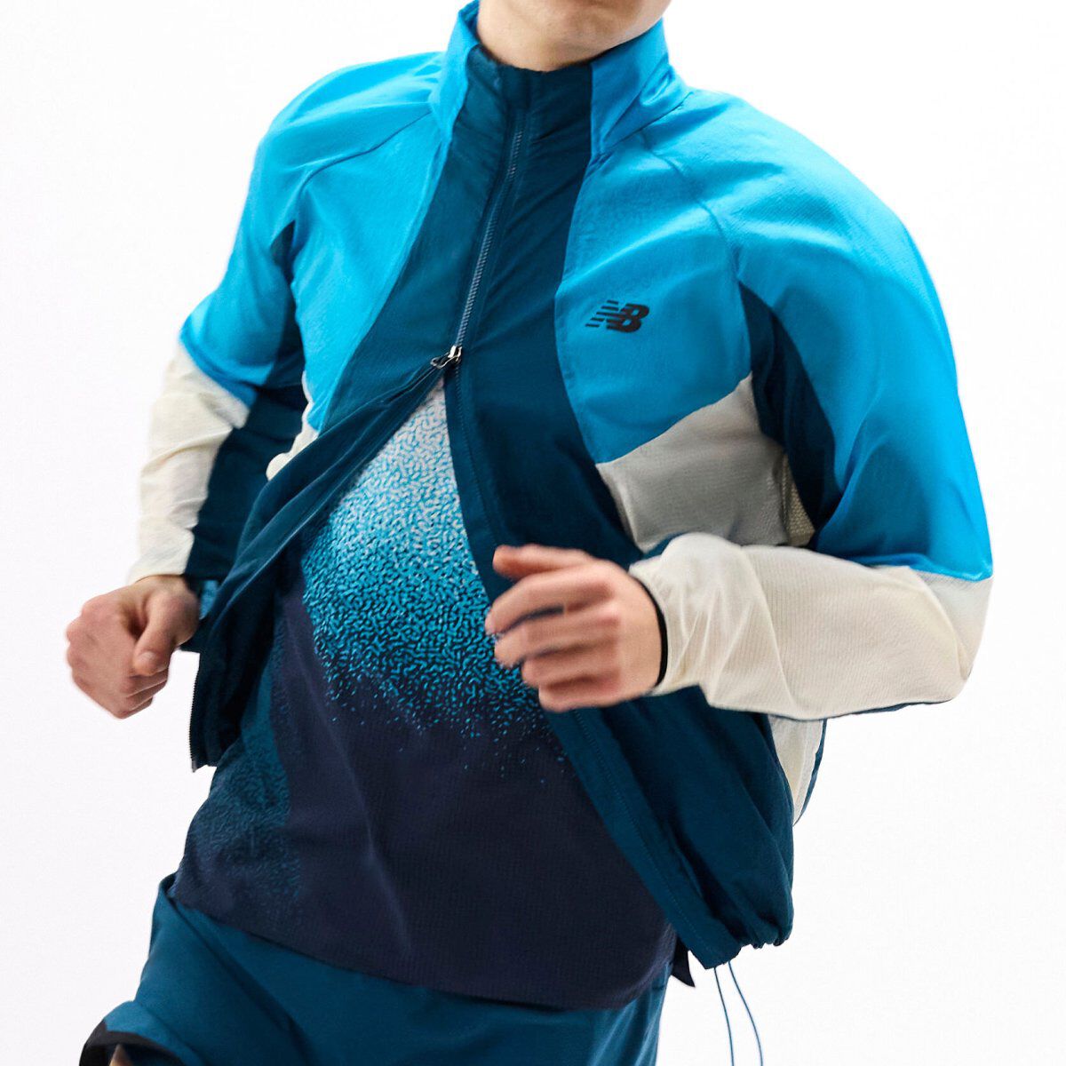 Close-up of a runner wearing a blue color-blocked jacket in motion, partially unzipped to reveal a patterned performance shirt against a white background. 