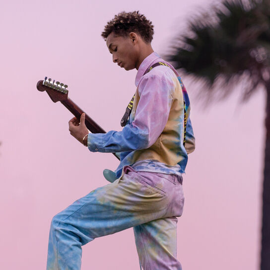 Jaden Smith porte une tenue tie-dye et joue de la guitare en plein air sur un fond rose.