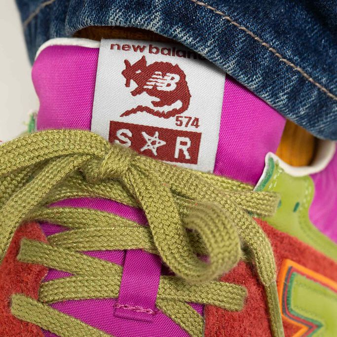 Gros plan du détail du dessus d'une chaussure de sport rouge NB x Stray Rats 574 aux accents roses et verts, présentant la marque NB et Stray Rats sur l'étiquette de languette