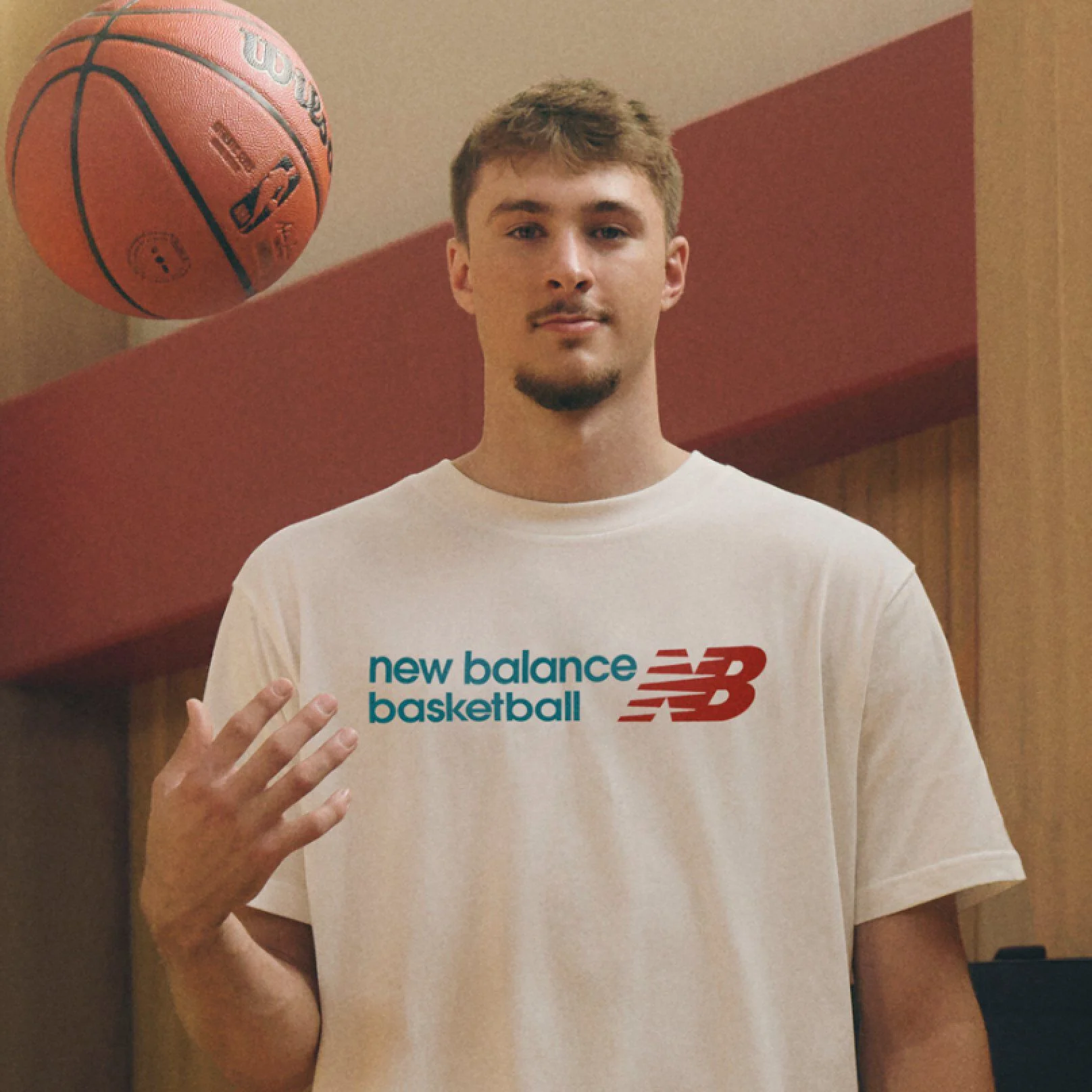 Une personne portant un t-shirt blanc &laquo; new balance basketball &raquo; lance un ballon de basket en l&rsquo;air d&rsquo;une main, debout &agrave; l&rsquo;int&eacute;rieur devant un fond en bois aux tons chauds et rouge.