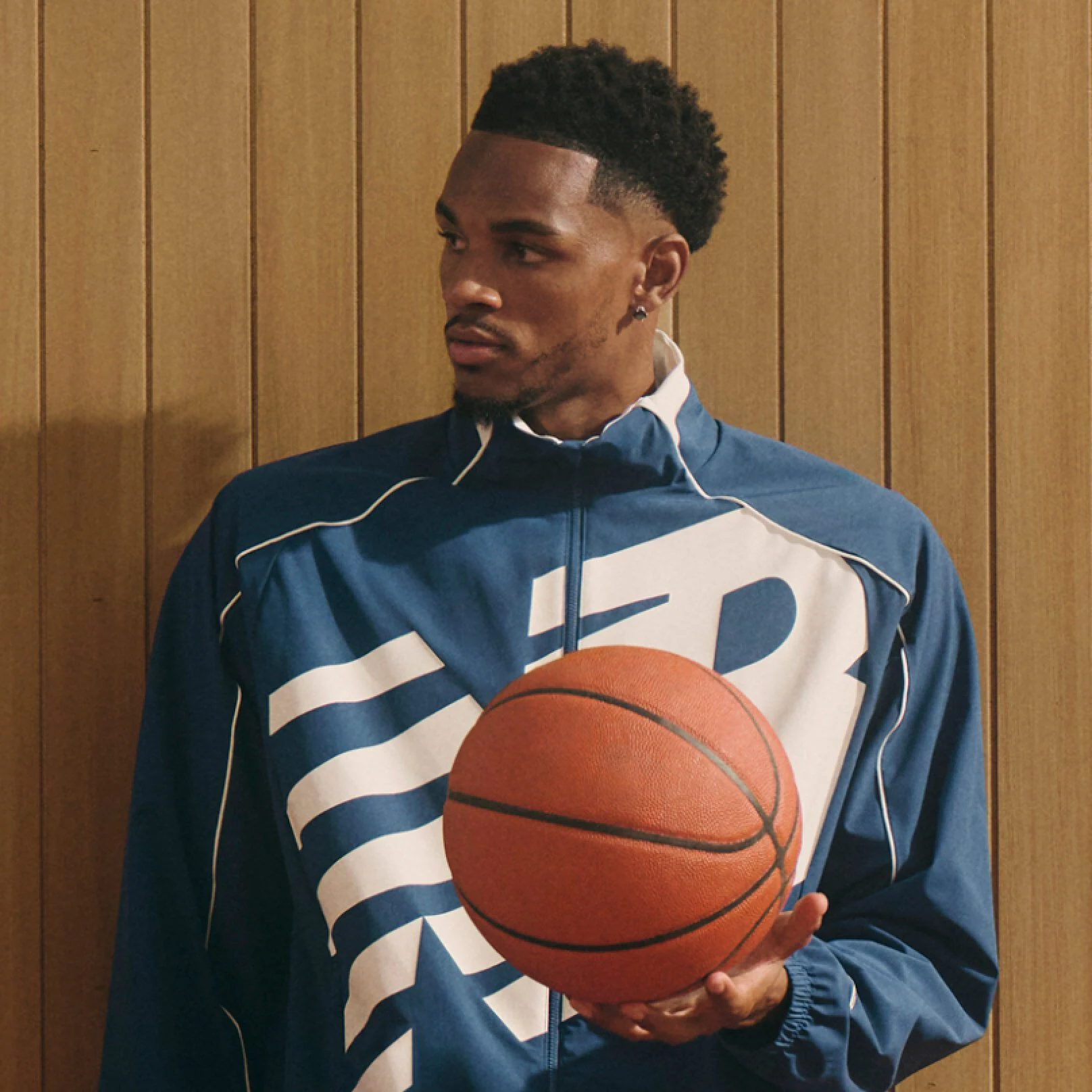 Une personne portant une veste bleue New Balance avec un grand logo blanc tient un ballon de basket devant un fond en panneaux de bois.