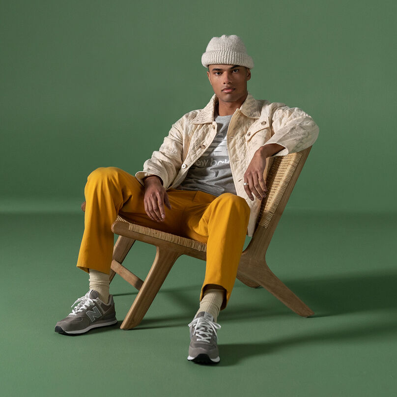 Man wearing 574 shoes and street clothes sitting on wooden chair against green studio background