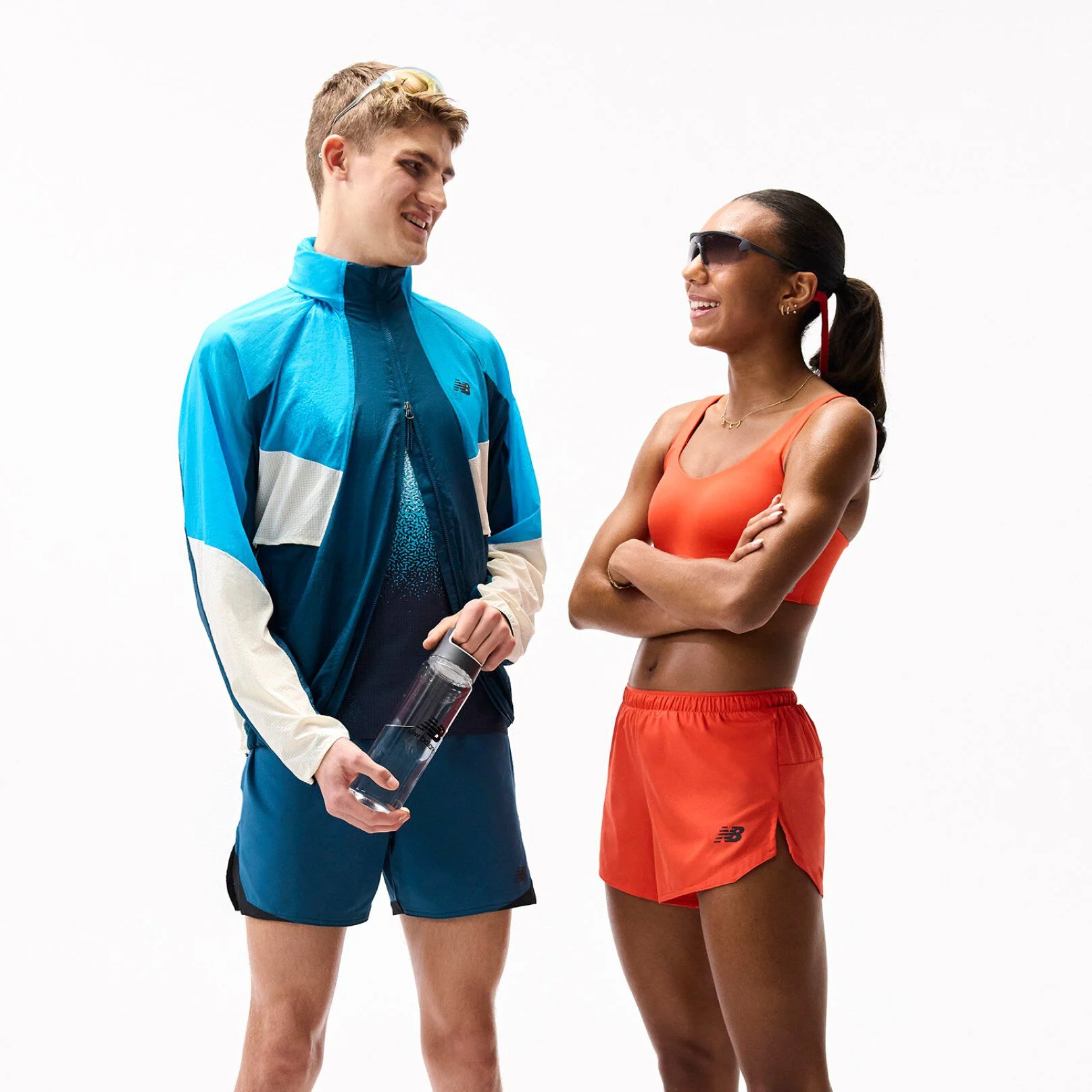 Two people in colorful athletic outfits stand facing each other on a plain white background, one holding a water bottle, both wearing sporty accessories and appearing mid-conversation. 