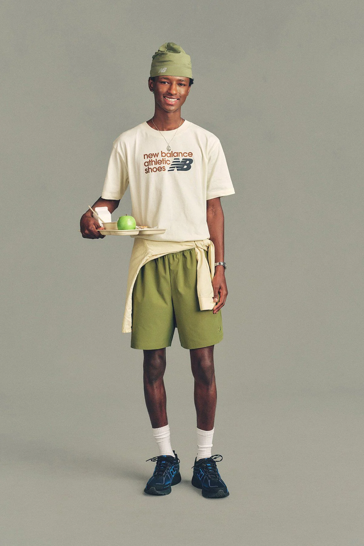 A man in a hat wearing a New Balance shirt and shorts standing in front of a grey studio background.