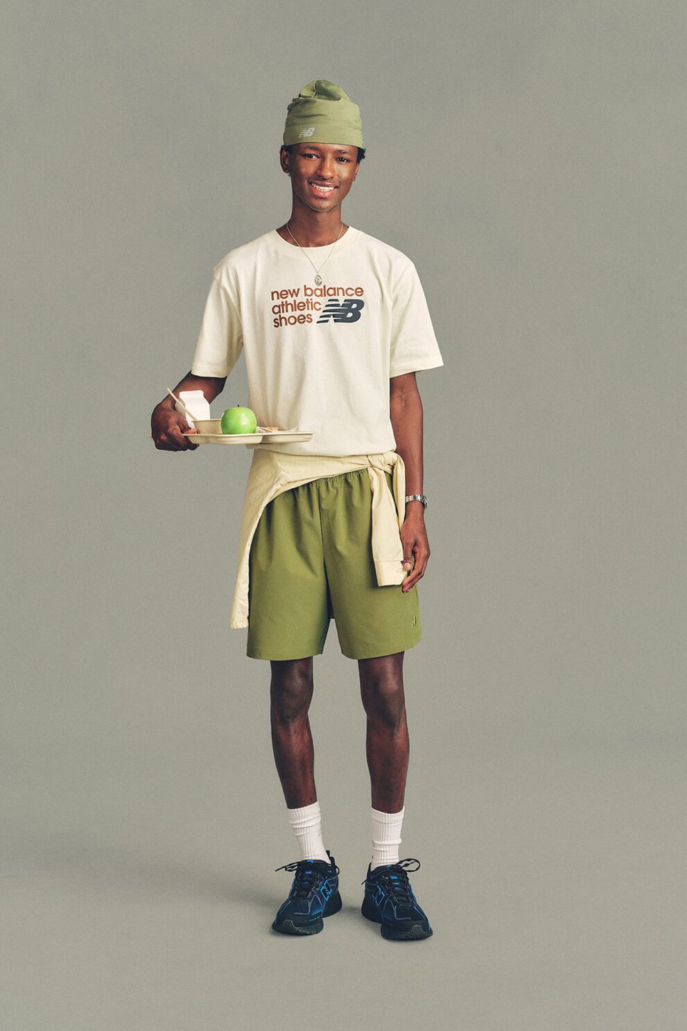 A man in a hat wearing a New Balance shirt and shorts standing in front of a grey studio background.