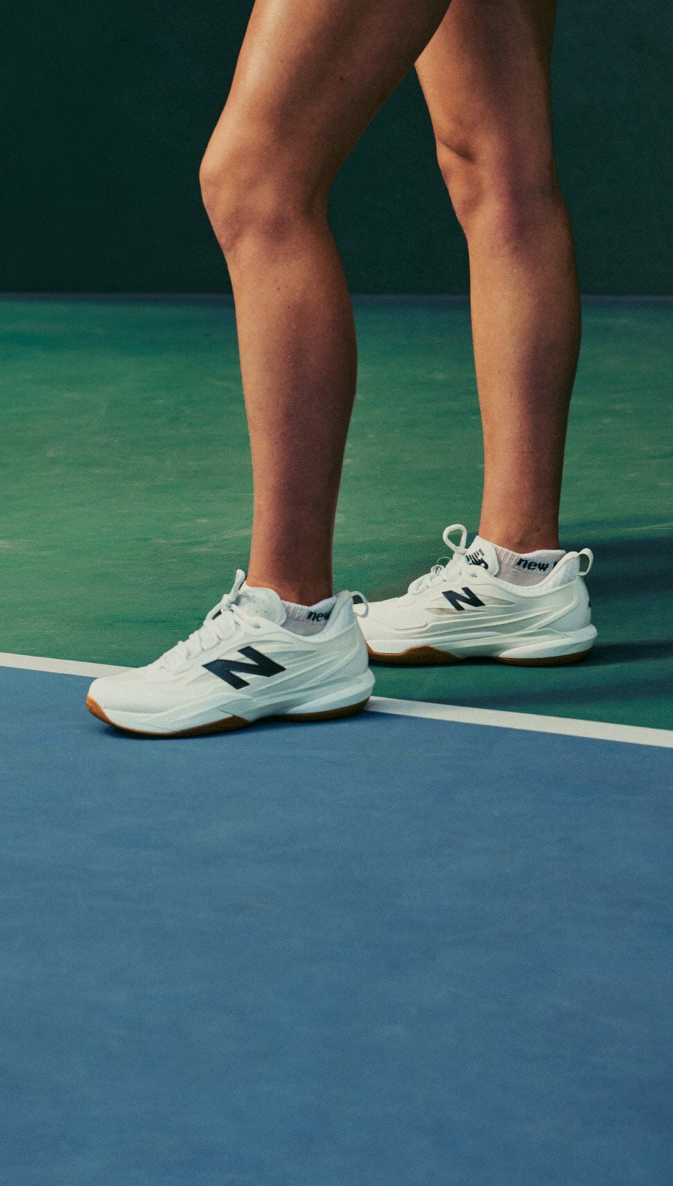 Gros plan de chaussures de tennis New Balance blanches port&eacute;es au bord du court sur un court int&eacute;rieur bleu et vert.