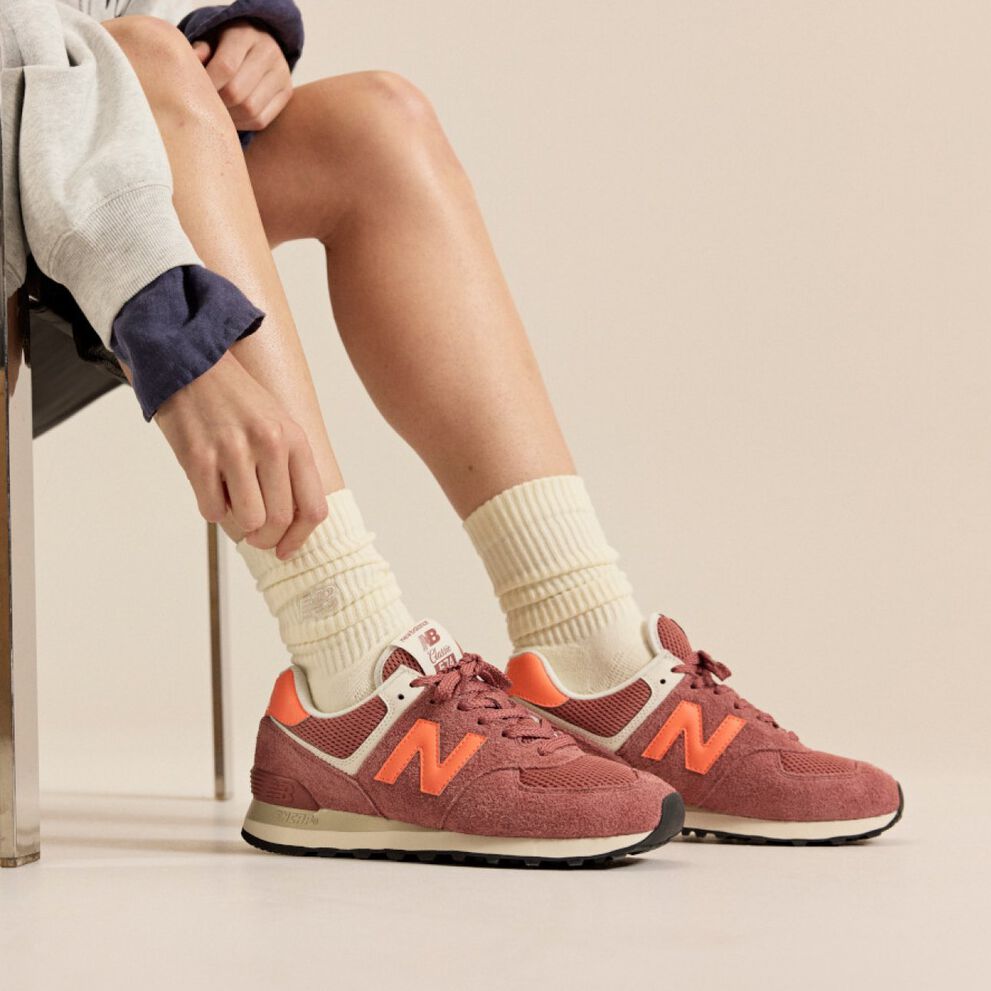 Personne assise sur une chaise portant des baskets New Balance rouge brique avec des accents orange et des chaussettes cr&egrave;me sur un fond neutre.