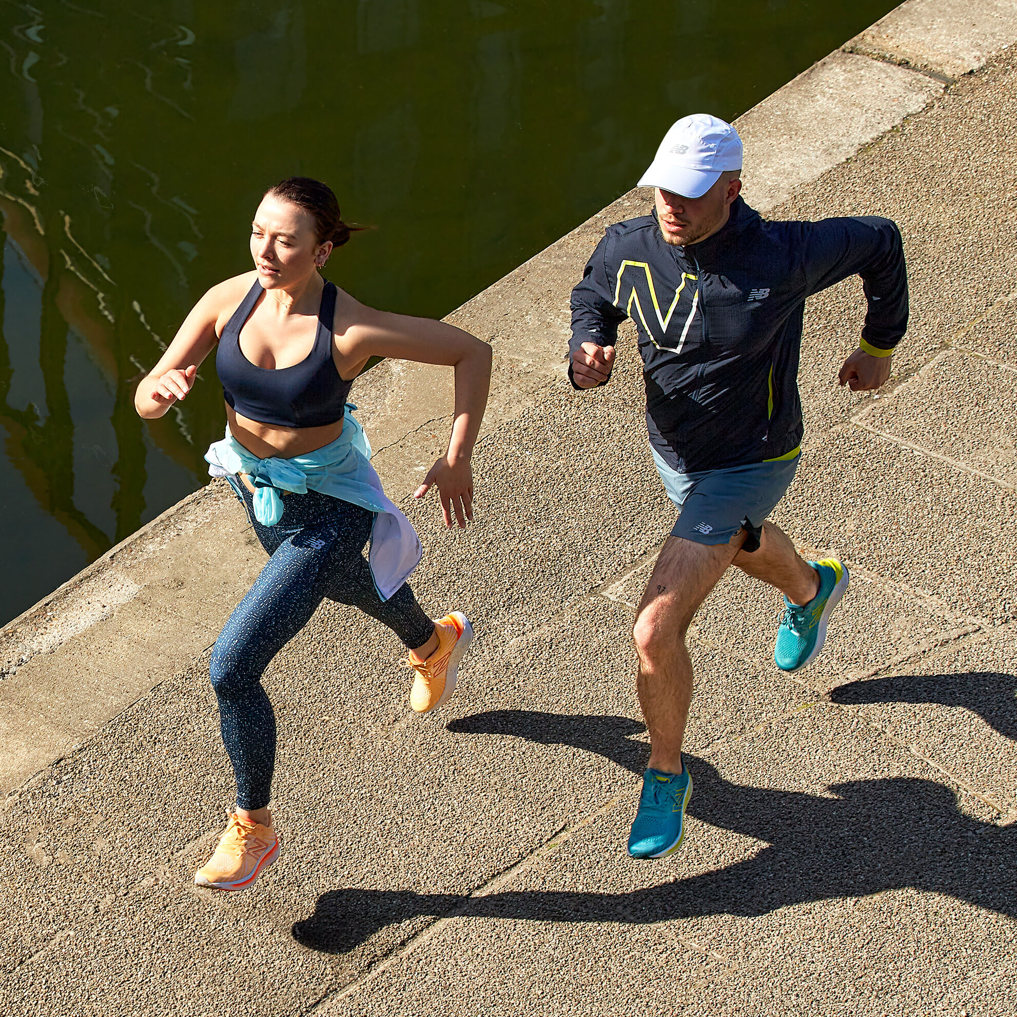 A male runner wearing a jacket and shorts and female runner wearing a running bra and leggings on concrete path next to water