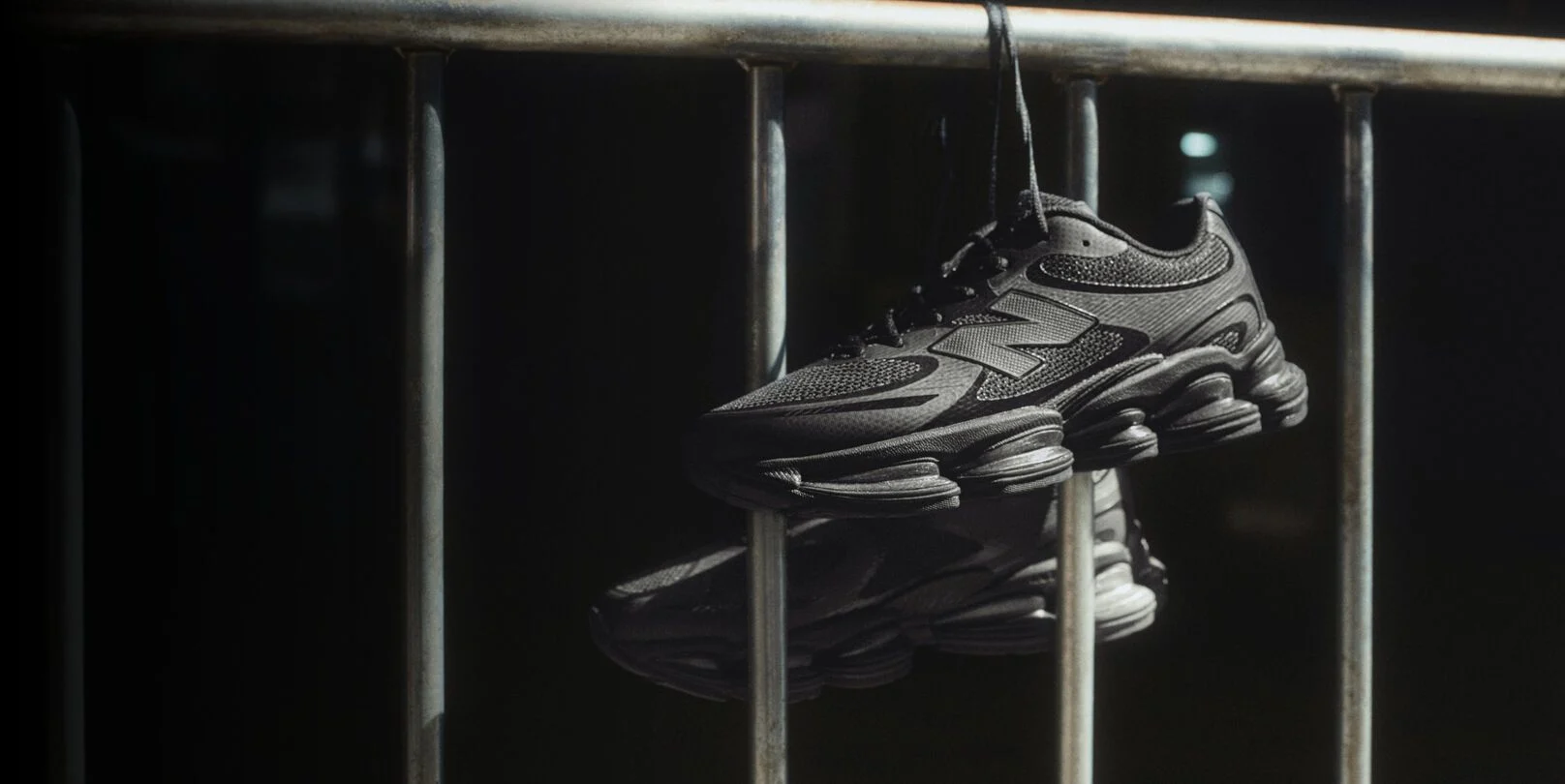 Black New Balance sneakers hanging by laces over a metal railing in dramatic lighting.