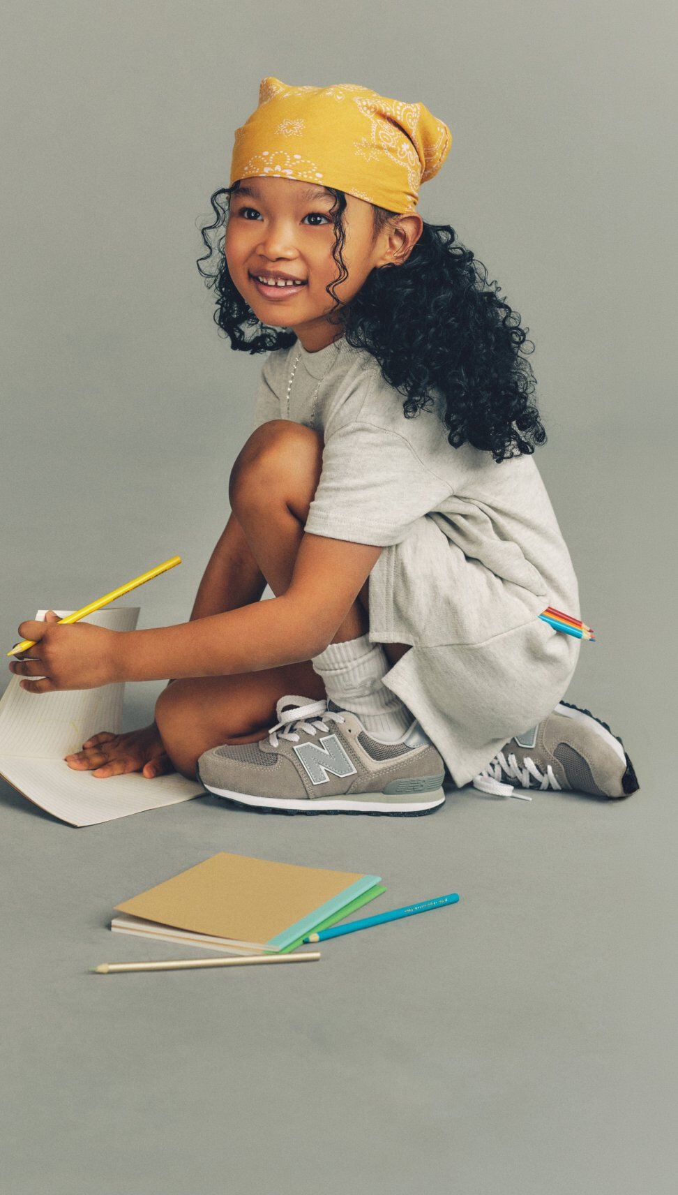 Child crouching on the floor writing in a notebook with colored pencils, wearing a yellow bandana, gray outfit, white socks, and gray New Balance sneakers against a plain background.