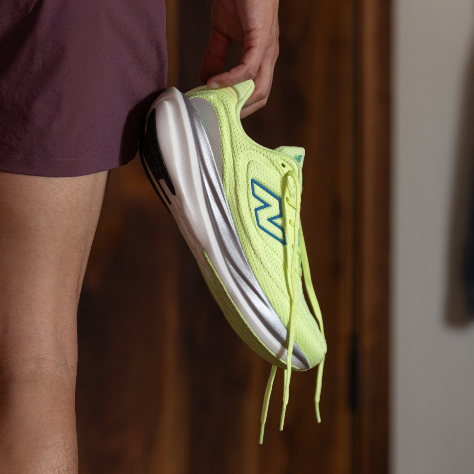 Gros plan d&rsquo;une chaussure de course New Balance jaune-vert vif avec logo bleu et semelle blanche, tenue par une personne portant un short bordeaux.&nbsp;