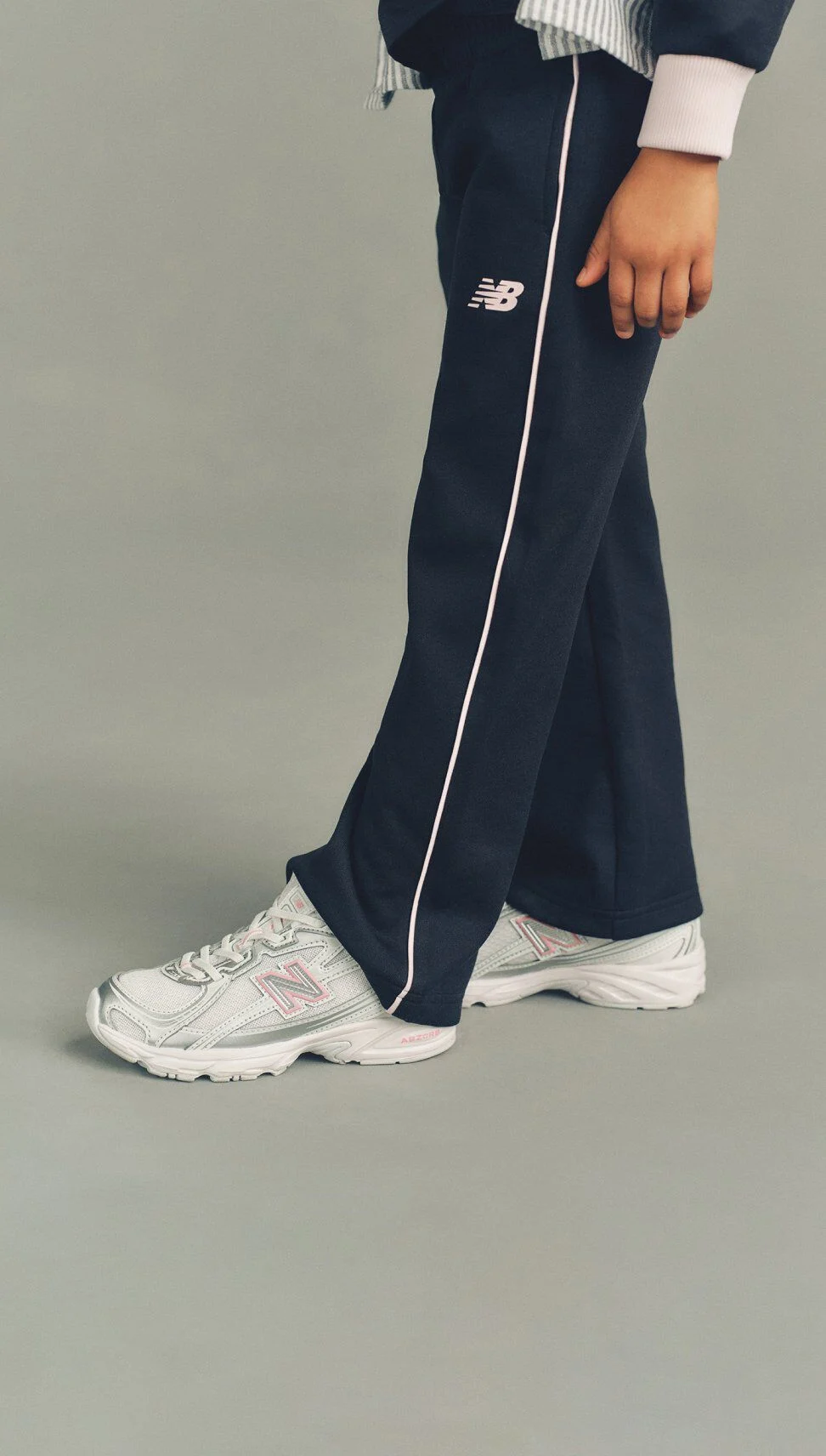 Close-up of a person wearing navy track pants with white piping and white New Balance sneakers with pink accents, standing against a neutral background.