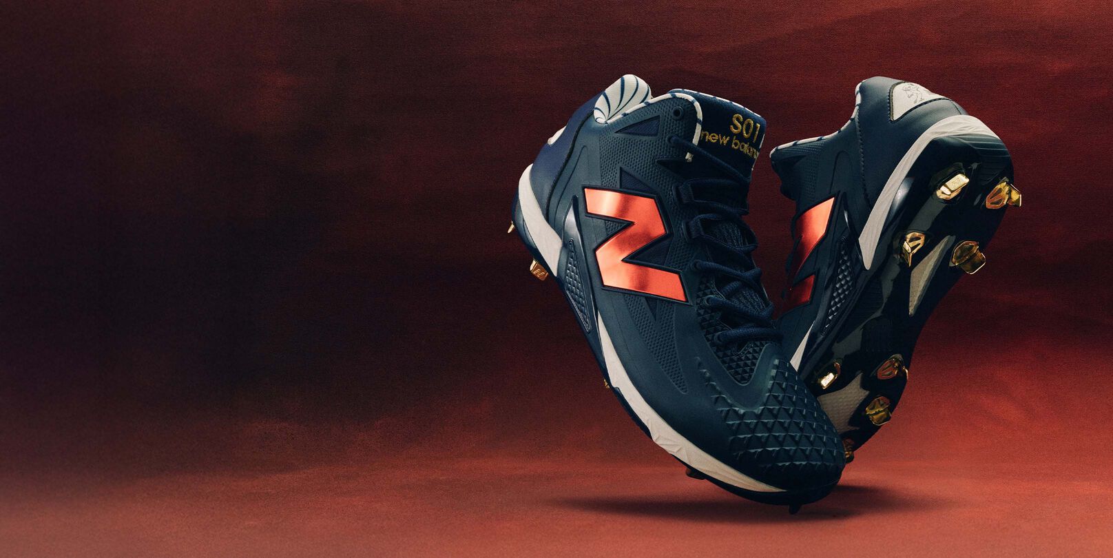 Pair of navy blue baseball cleats with metallic red New Balance logos and gold studs, posed against a warm red background.