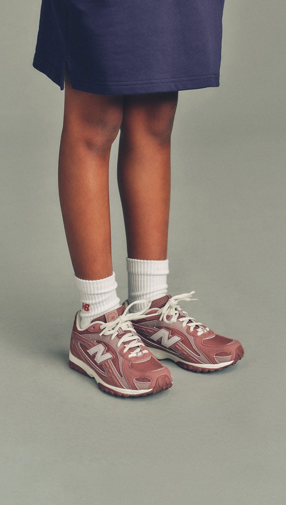 Close-up of a child wearing white socks and pink New Balance sneakers standing against a neutral background.