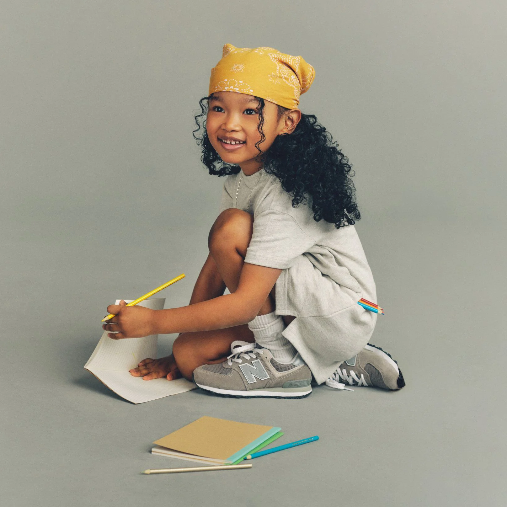 Child crouching on the floor writing in a notebook with colored pencils, wearing a yellow bandana, gray outfit, white socks, and gray New Balance sneakers against a plain background.
