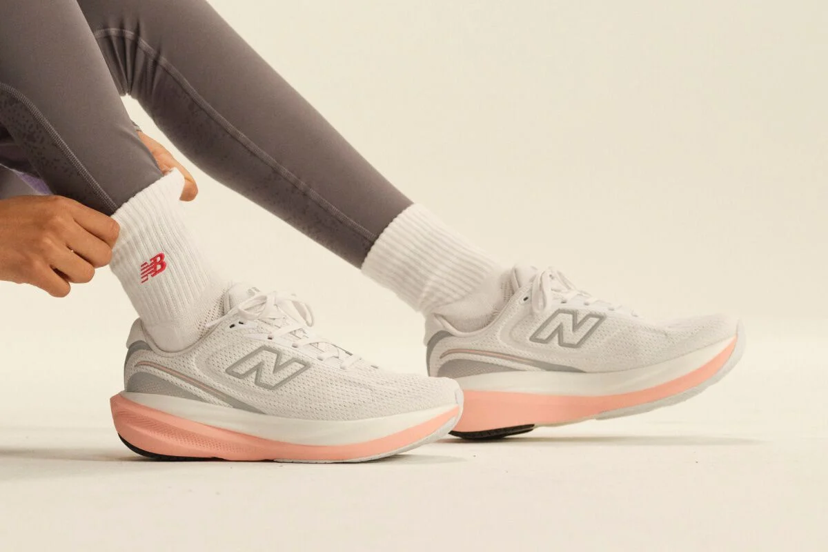 Close-up of white New Balance running shoes with pink midsoles paired with white crew socks and gray leggings in a clean studio setting.