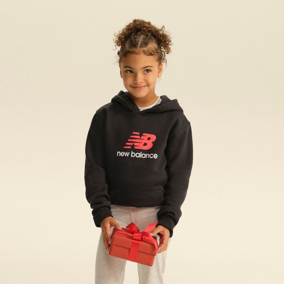 Child wearing a black New Balance hoodie and gray pants holding a red gift box against a light studio backdrop. 
