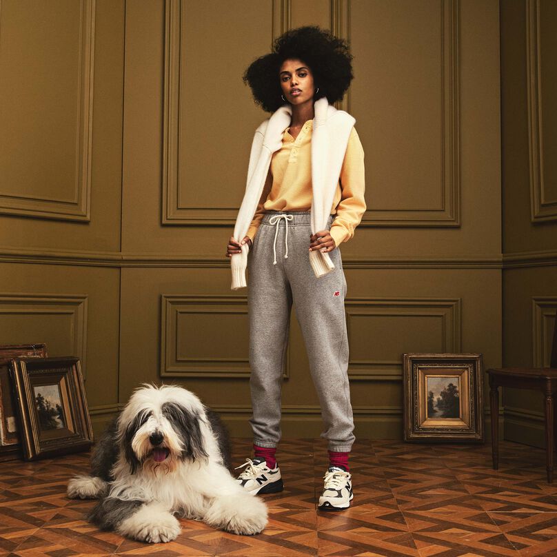 Femme debout dans un bel environnement intérieur et portant un pantalon de survêtement et des chaussures MADE in USA. Un grand chien est couché à côté d’elle.