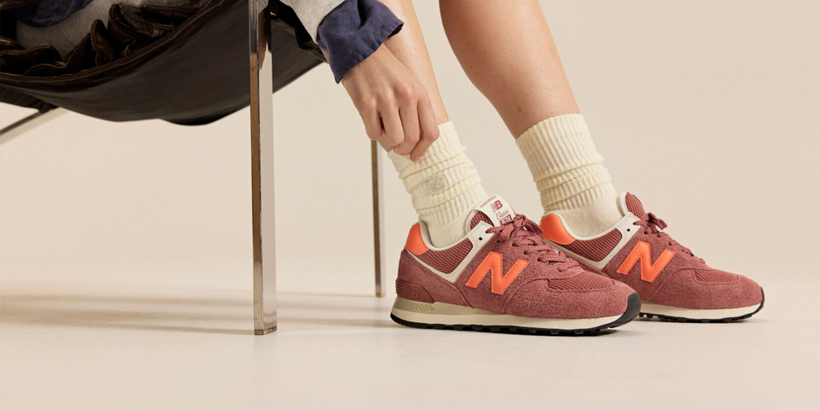 Personne assise sur une chaise portant des baskets New Balance rouge brique avec des accents orange et des chaussettes cr&egrave;me sur un fond neutre.