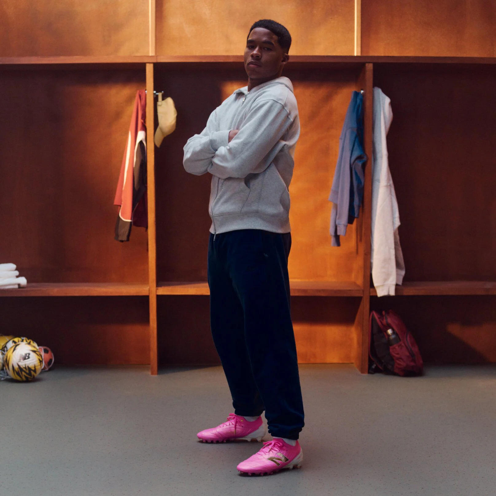Endrick standing in a wooden locker room with arms crossed, wearing a light gray zip‑up hoodie, dark athletic pants, and bright pink soccer cleats; open lockers behind display hanging apparel, folded clothing, a backpack, and soccer balls on the floor.