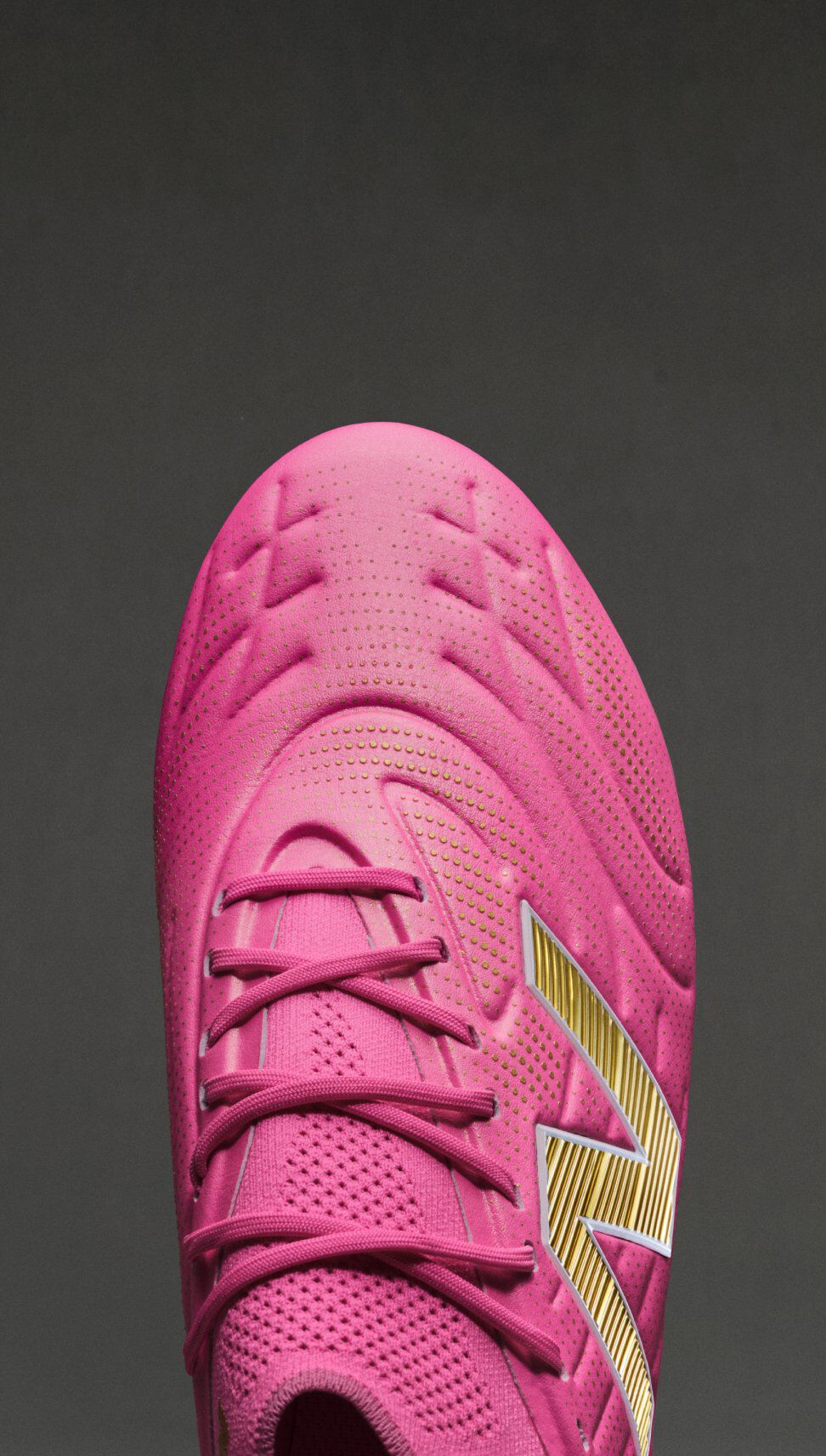 Top-down close-up of a bright pink New Balance soccer cleat on a dark gray background, showing textured upper detailing, pink laces, and a metallic gold &ldquo;N&rdquo; logo along the side.