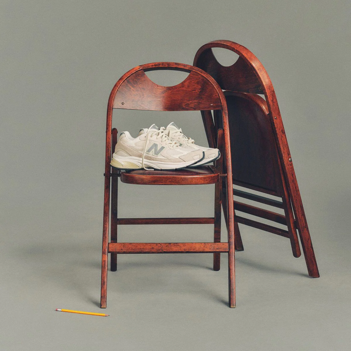 Image studio d&rsquo;une paire de chaussures de sport New Balance couleur cr&egrave;me plac&eacute;e sur une chaise pliante en bois fonc&eacute;, avec une deuxi&egrave;me chaise assortie repli&eacute;e et appuy&eacute;e derri&egrave;re.