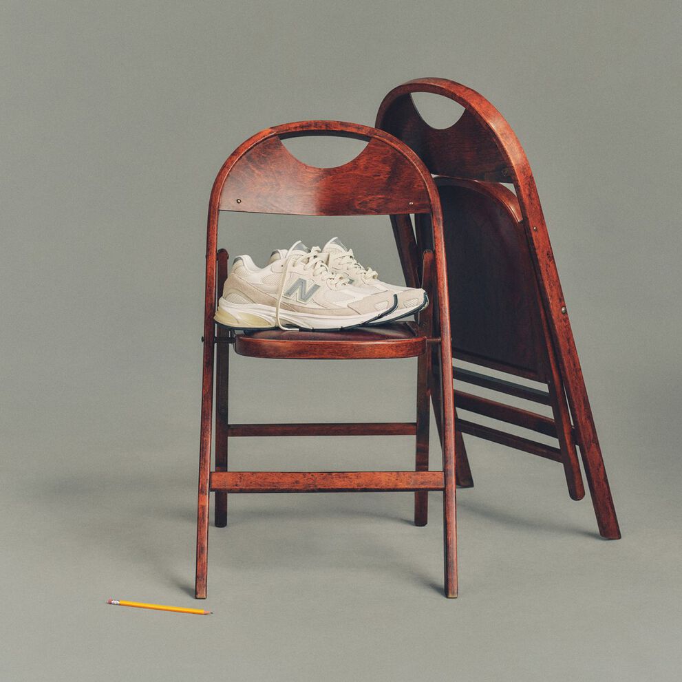 Image studio d&rsquo;une paire de chaussures de sport New Balance couleur cr&egrave;me plac&eacute;e sur une chaise pliante en bois fonc&eacute;, avec une deuxi&egrave;me chaise assortie repli&eacute;e et appuy&eacute;e derri&egrave;re.