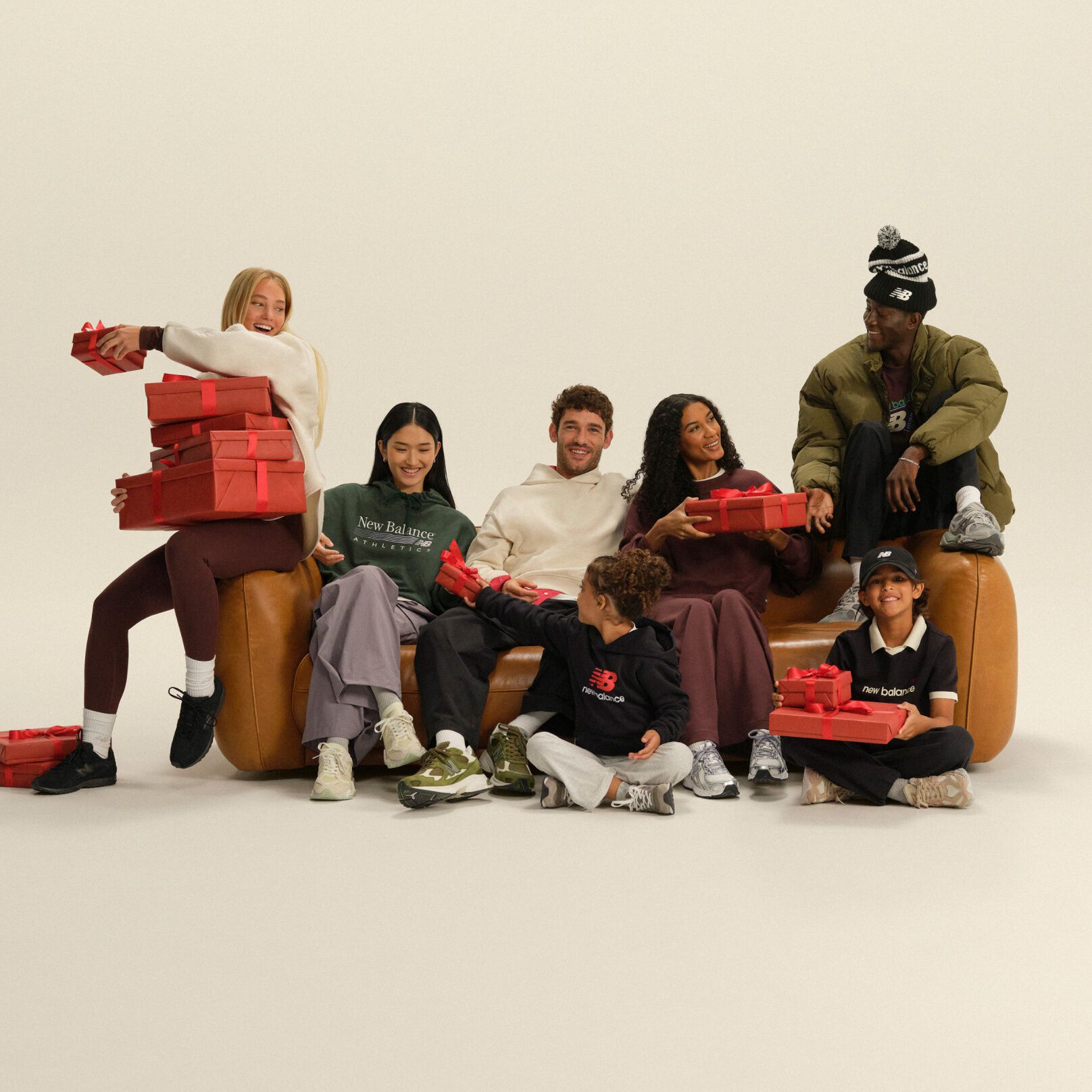 Group of adults and children in casual athletic apparel gathered around a sofa, exchanging red gift boxes in a festive studio setting.