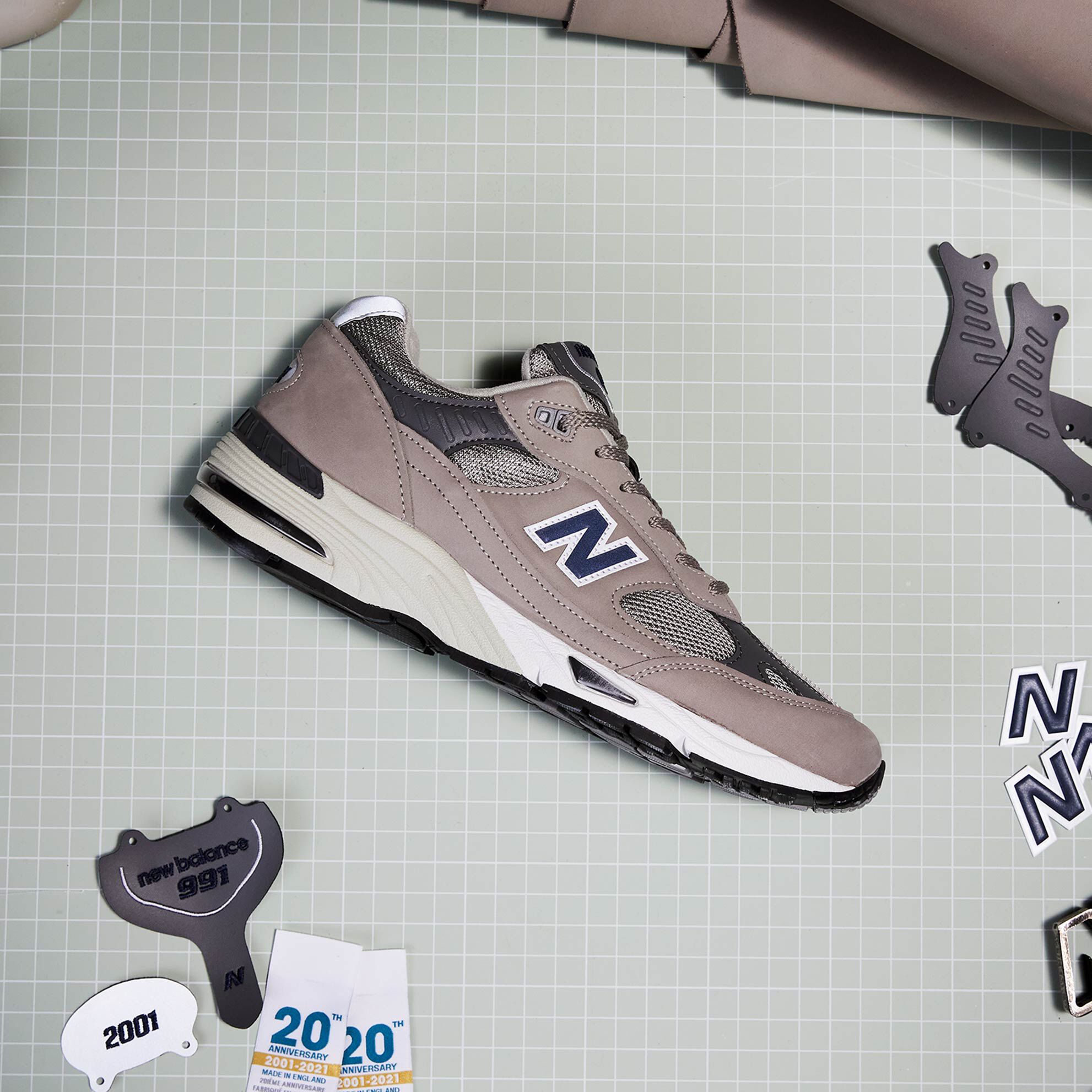 A men’s grey NB 991 sneaker sitting on a grid-lined cutting mat amongst the parts and pieces from which it was constructed