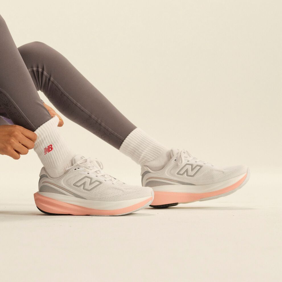 Close-up of white New Balance running shoes with pink midsoles paired with white crew socks and gray leggings in a clean studio setting.