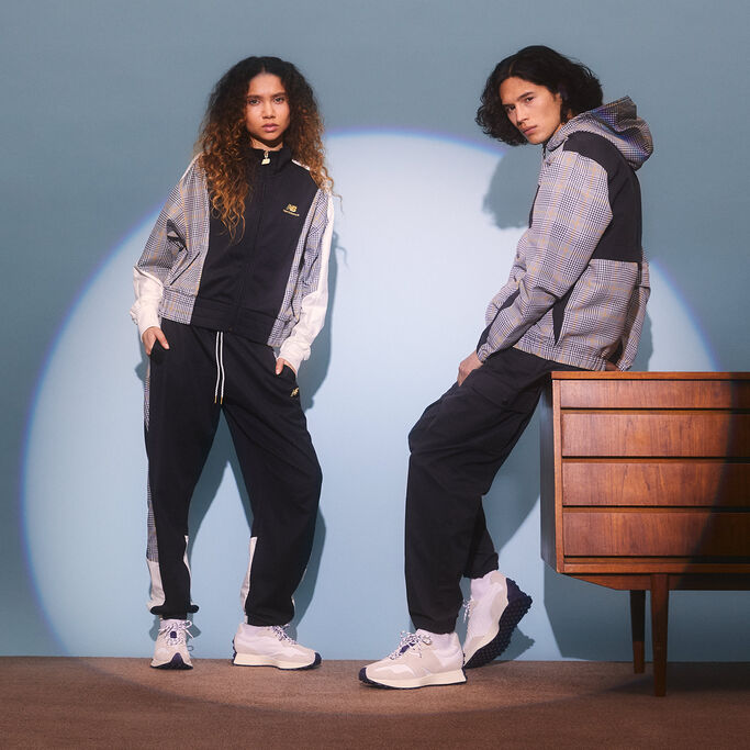 Un homme et une femme posent en tenue NB Athletics et en chaussures 327 dans un studio avec du mobilier et de la moquette vintage.