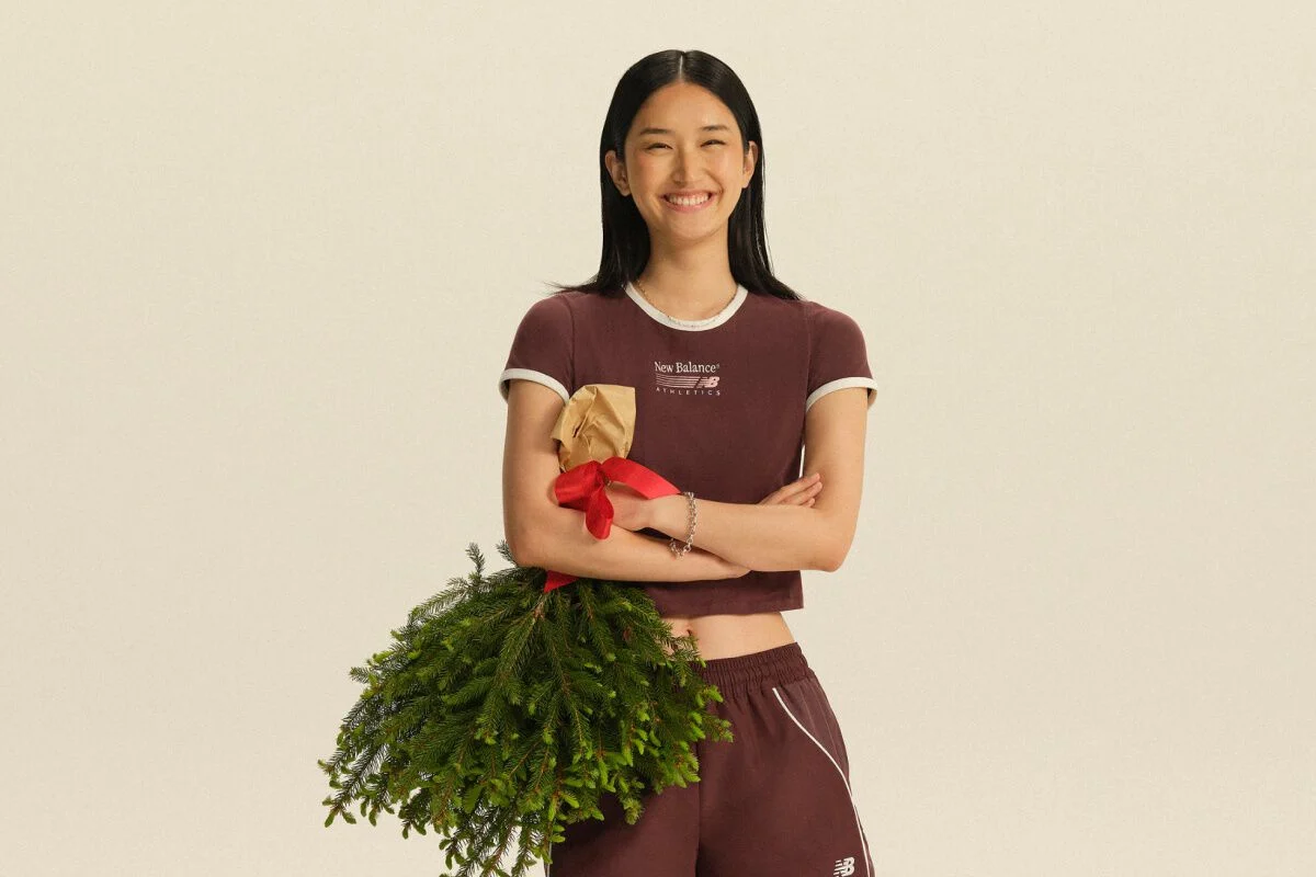 Person wearing a burgundy New Balance athletic set holding a wrapped gift and evergreen branches against a neutral studio background. 