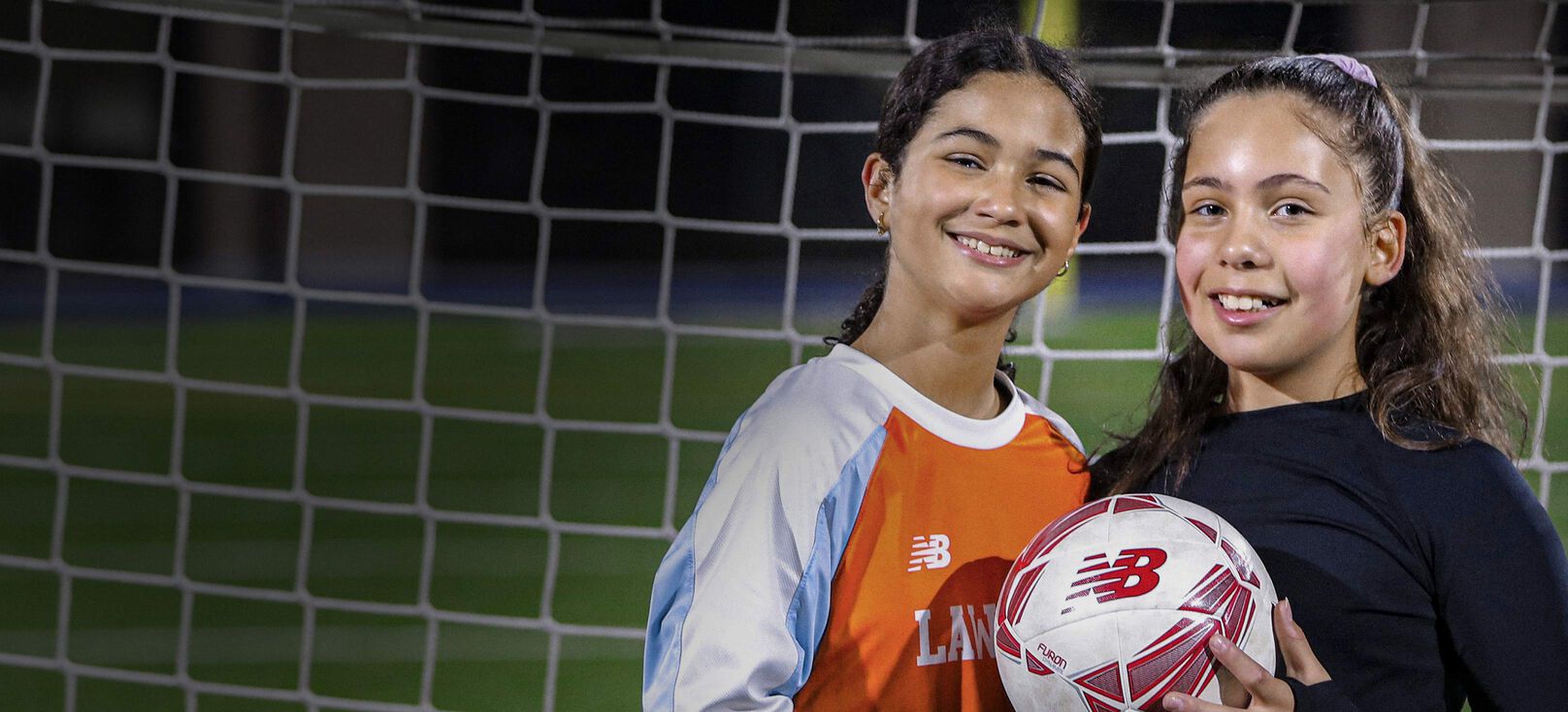 Deux adolescentes souriantes posant avec un ballon de football devant un but.